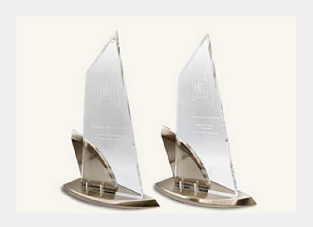Two clear glass awards with metal bases are displayed side by side against a plain light background. The awards are triangular with engraved text and a logo visible on each.