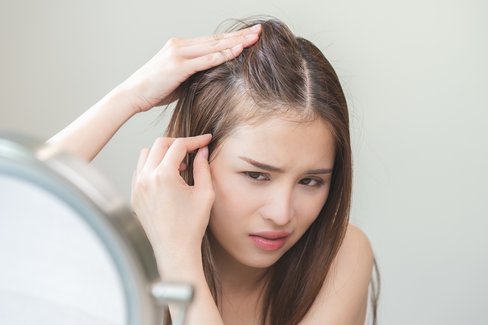 A woman examines her scalp in the mirror, parting her hair and looking concerned, with her fingers lifting her hair near the hairline, searching for signs of Hair Loss or Telogen Effluvium.