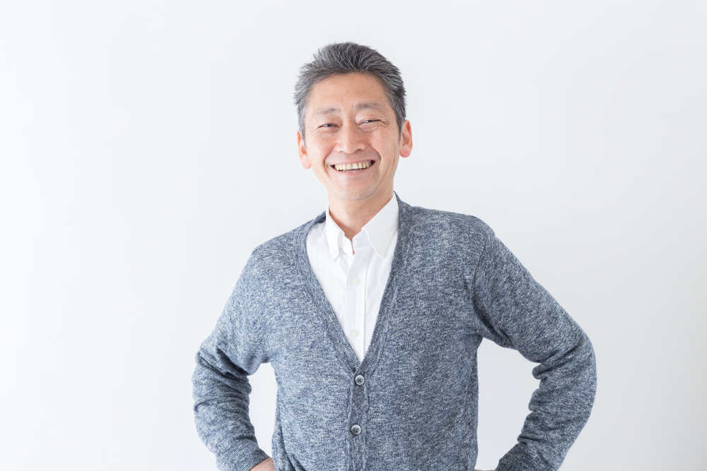 Middle-aged man with gray hair smiling, standing with hands on hips, wearing a white shirt and a gray cardigan, against a plain white background—an image that challenges common grey hair myths and invites conversation about the real causes of grey hair.