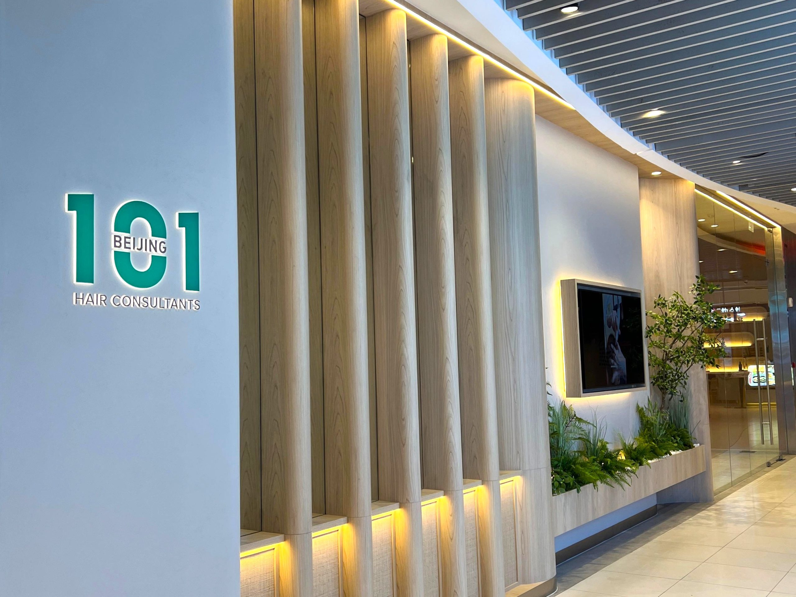 Modern clinic entrance showcasing 101 BEIJING HAIR CONSULTANTS—a credible brand—with vertical wooden panels, a wall-mounted TV, green plants, and bright indoor lighting reflecting strong branding.