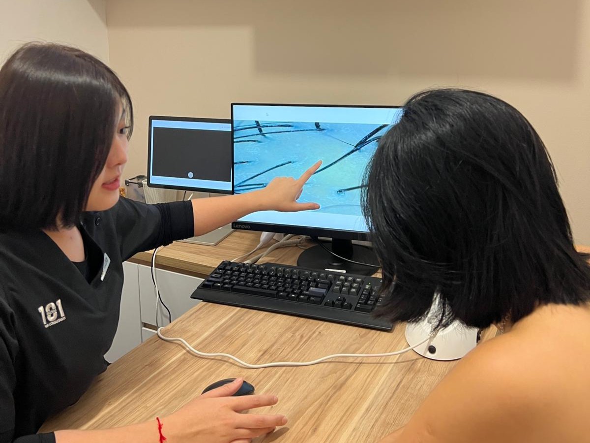 A woman points to a close-up image of hair on a computer screen while discussing it with another person at a wooden desk.