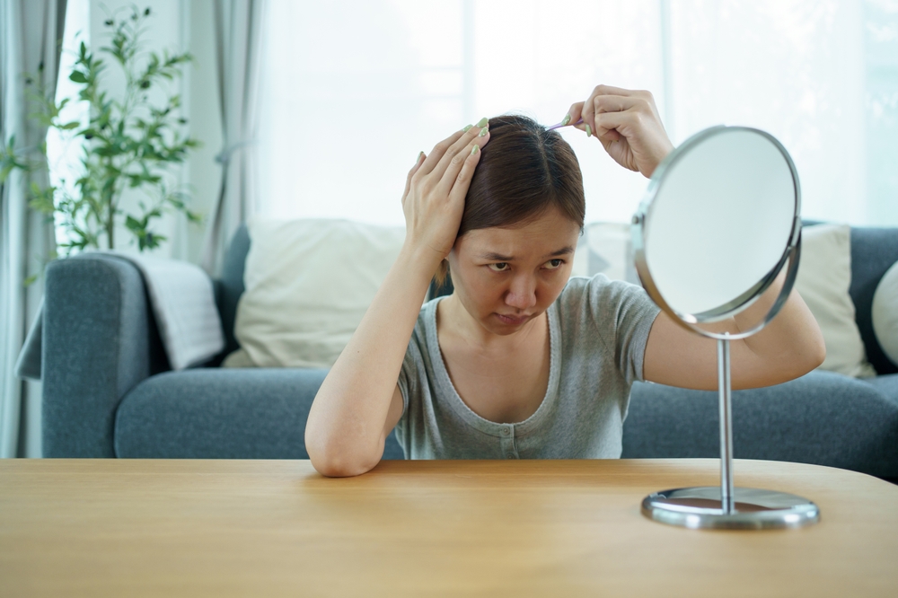 A woman sits at a table in front of a mirror, parting her hair with her hands and examining her scalp, looking concerned—perhaps wondering about grey hair myths like “does plucking cause more?” A sofa and plants are visible in the background.