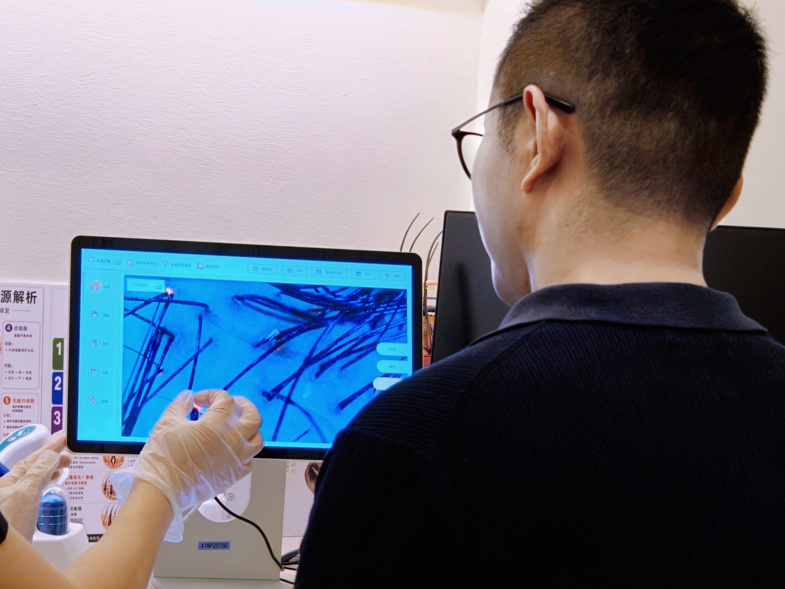 During a hair consultation, a person in gloves points at a computer screen showing a magnified blue and black image, while another observes, both focusing on what to expect during the first visit.