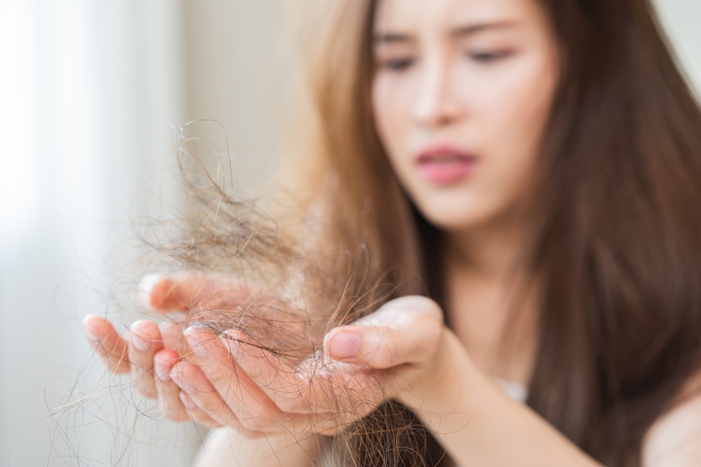 A woman with long brown hair looks worried while holding a clump of fallen hair in her hands, indicating concern about early signs of hair loss.