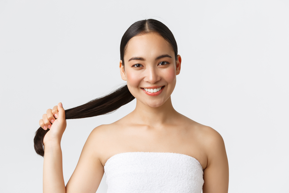 Smiling woman in a white towel holds her long dark hair to the side, showcasing the results of her recent hair treatment, standing against a plain light background.
